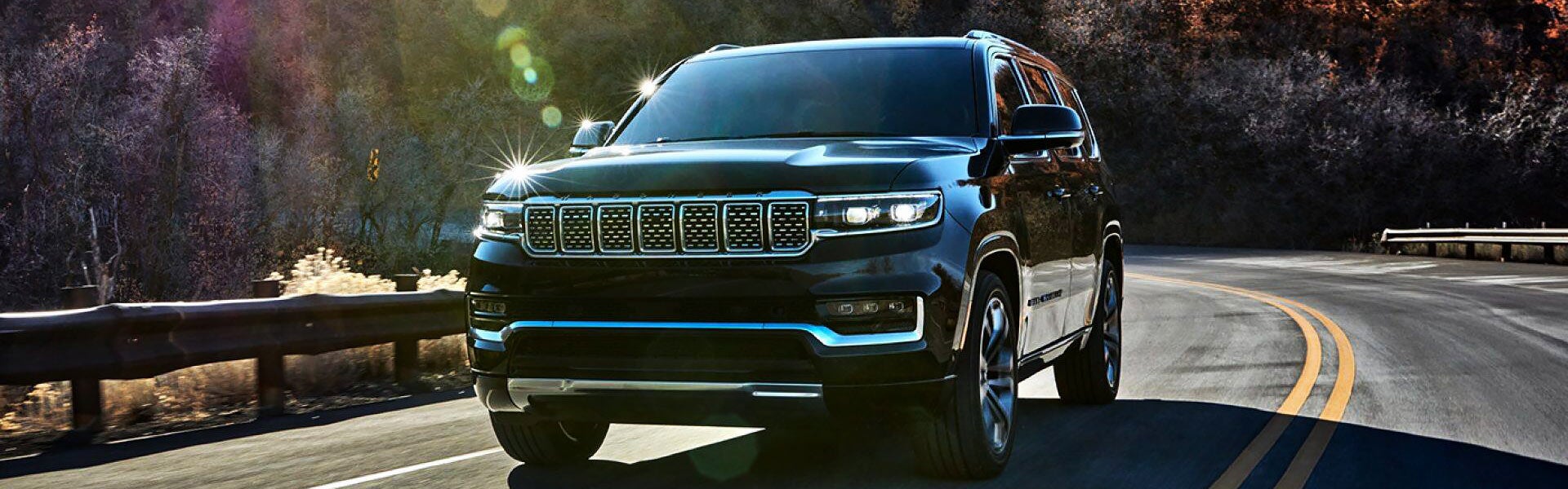 A shiny black SUV with bright headlights drives on a winding road with a metal guardrail and trees.