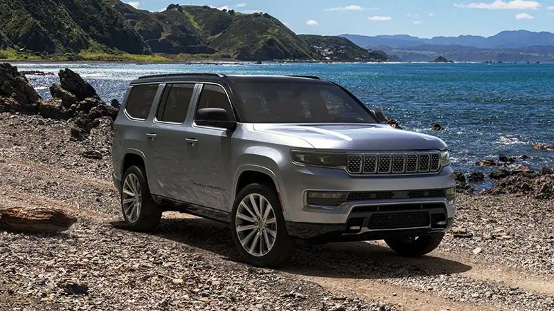 Silver Jeep Wagoneer SUV parked on a rocky shoreline next to a blue ocean, with mountains in the background.