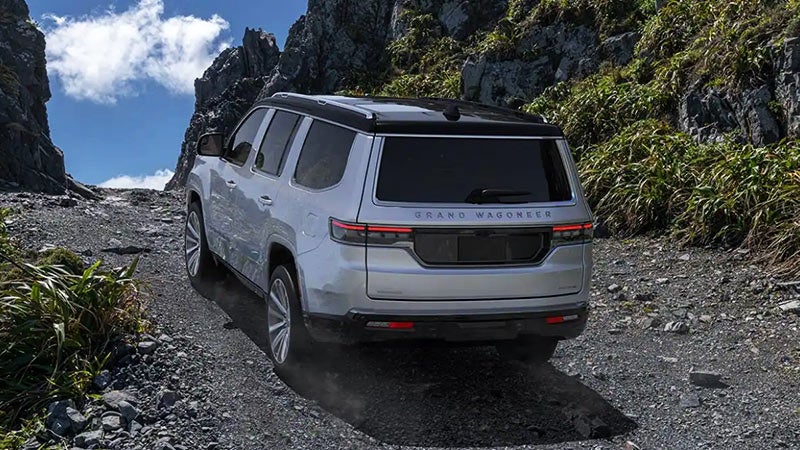 Silver Grand Wagoneer SUV drives aggressively up a rocky mountain pass under a blue sky.