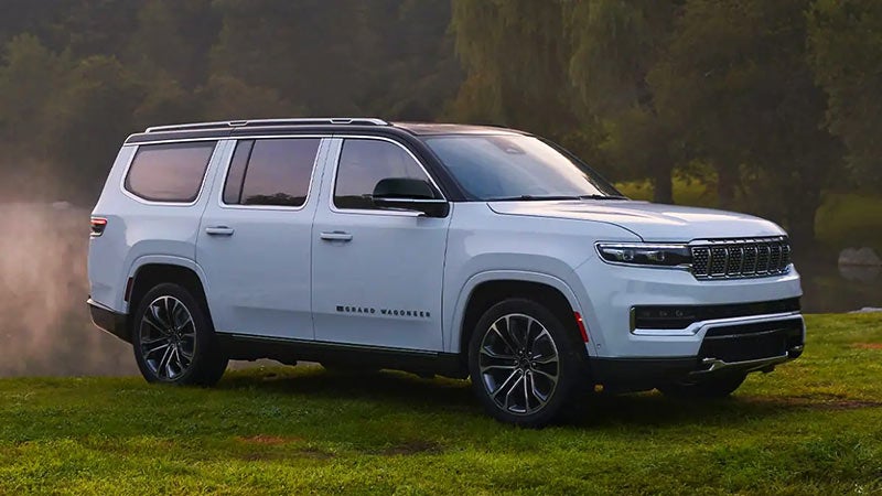 White 2022 Grand Wagoneer parked on a grassy hill with a forested background and a body of water.