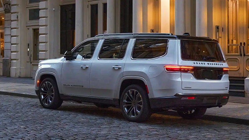 A white 2022 Grand Wagoneer SUV is parked on a cobblestone street in front of a building with lit windows.