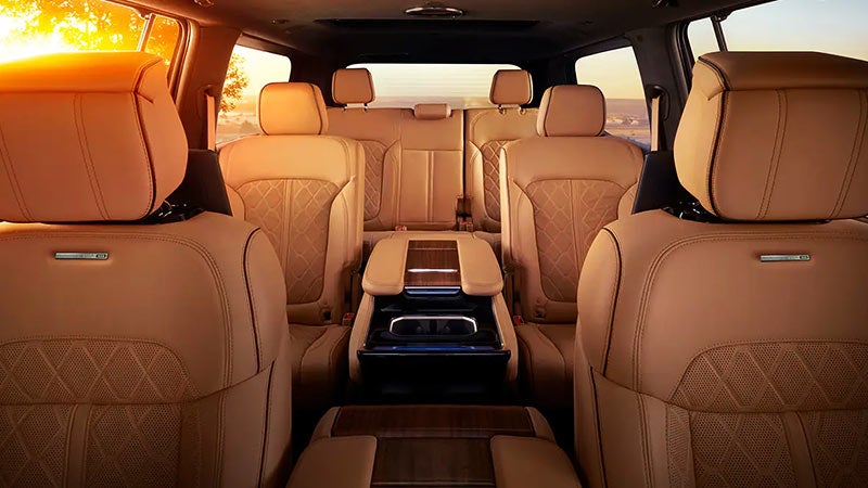 Interior of a large car with three rows of tan leather seats, centered by a wooden console. Sunlight streams through windows.