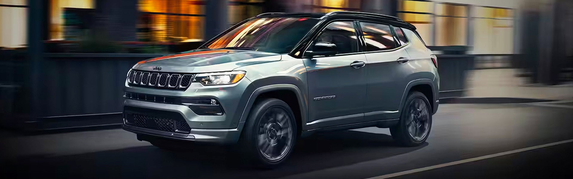 A gray 2024 Jeep Compass SUV drives left across a city street at high speed, blurred background.