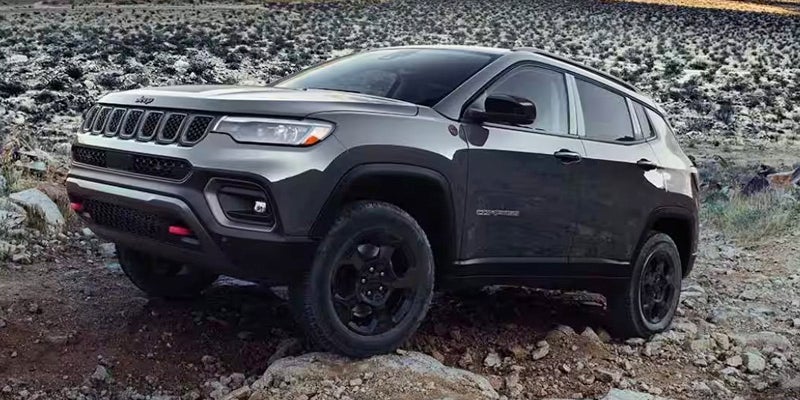A dark gray Jeep Compass Trailhawk SUV parked on a rocky, uneven dirt and gravel terrain.
