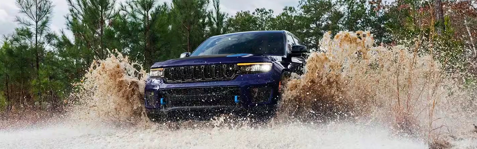A dark blue Jeep Grand Cherokee drives through deep muddy water, splashing mud and water high into the air.