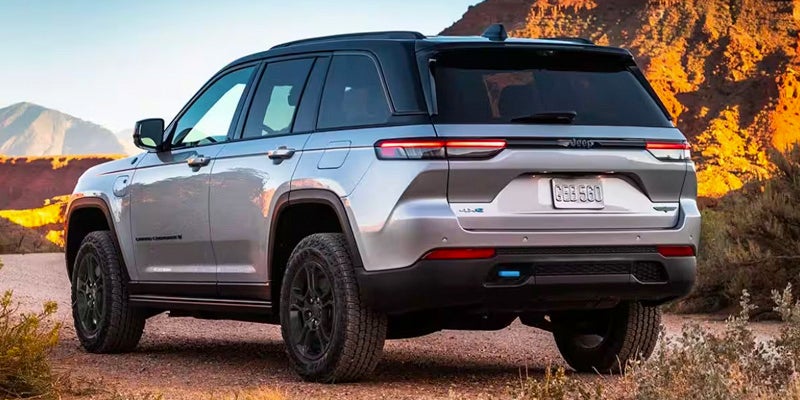 Rear quarter view of a silver and black Jeep Grand Cherokee 4xe parked on a dirt path with mountains in the background.