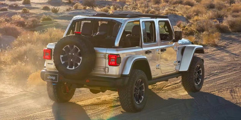 Silver Jeep Wrangler with a soft top traversing a dusty desert trail at sunset.
