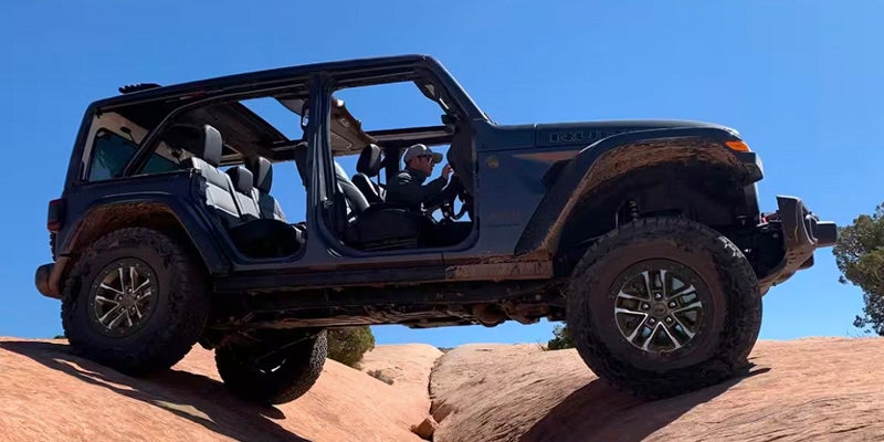 Dark blue Jeep Wrangler crossing a rocky crevice, driver visible, sunny blue sky.