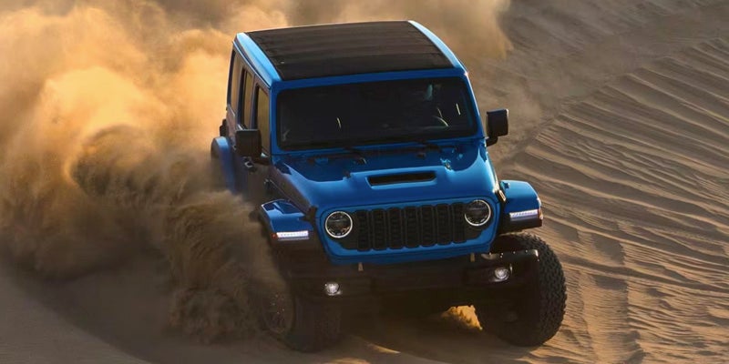 A blue-green Jeep Wrangler Rubicon kicks up billowing dust as it drives at an angle over a sand dune.