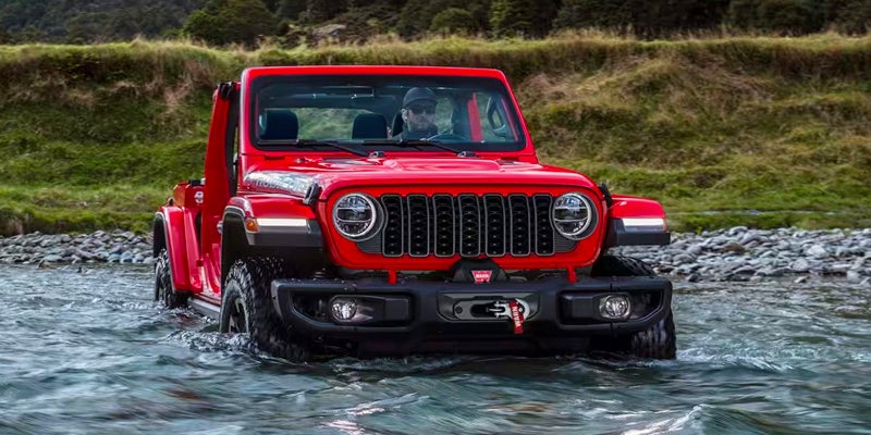A red Jeep Wrangler Rubicon 392 with a driver, fording a shallow river in a natural, green-banked setting.