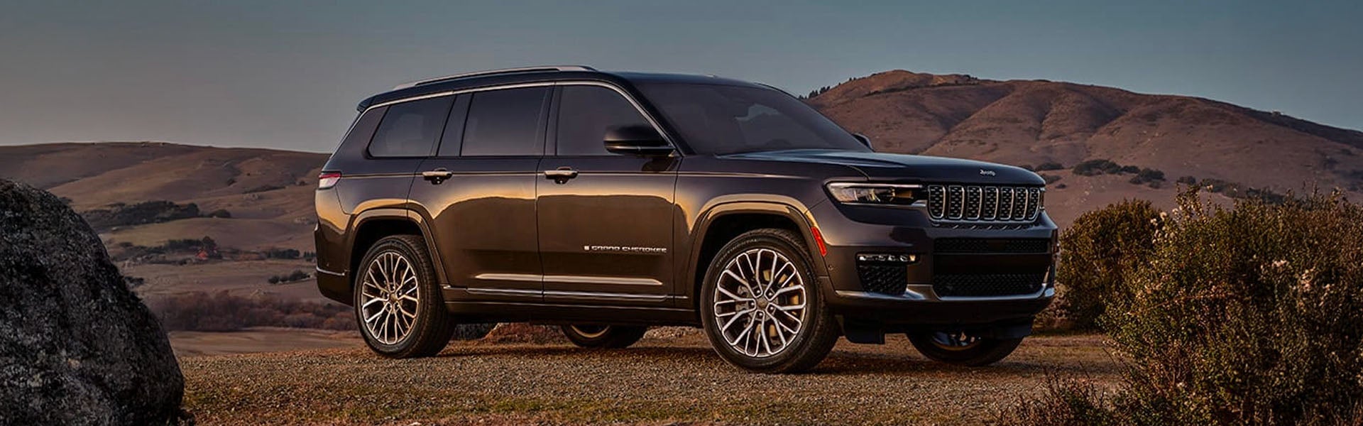 A dark gray Jeep Grand Cherokee L parked on a dirt hill with rolling brown hills and a blue sky in the background.