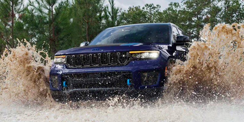 Blue Jeep Grand Cherokee 4xe splashing through muddy water with pine trees and green foliage in background.