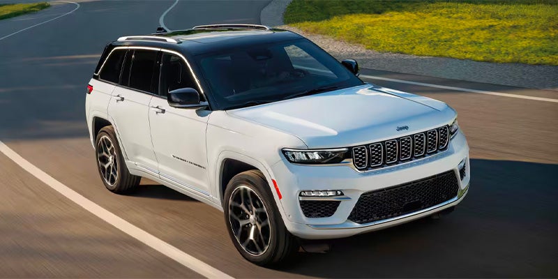 White Jeep Grand Cherokee L, with a black roof and silver roof rails, driving on a road with green grass.