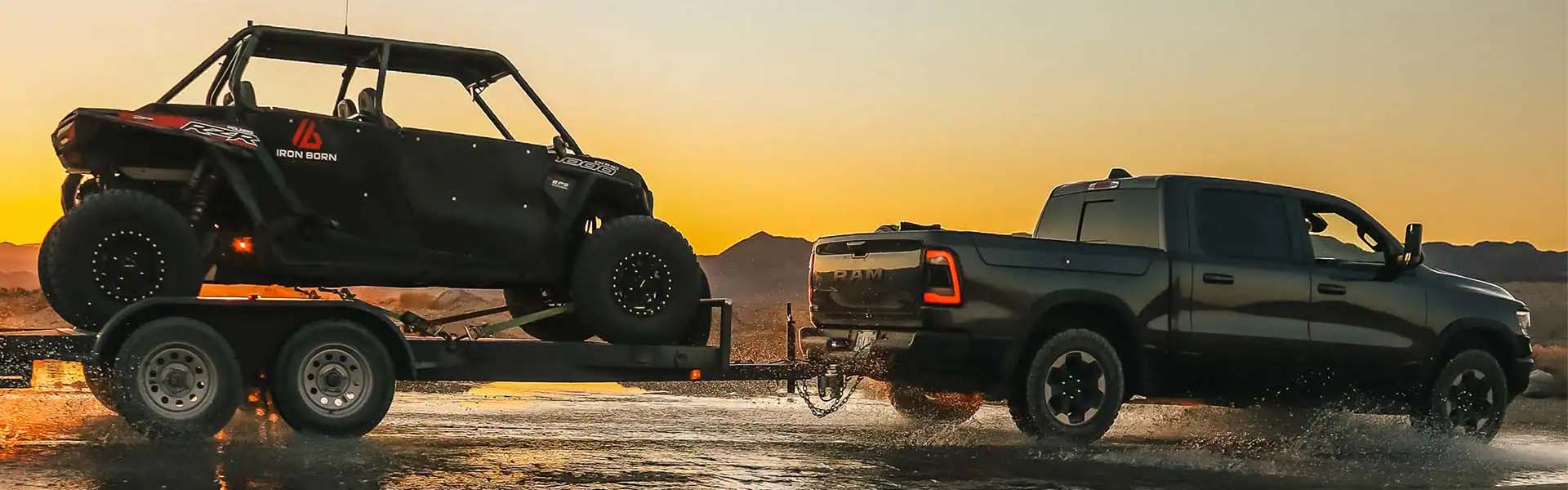 Black pickup truck towing a utility task vehicle (UTV) on a trailer through shallow water at sunset.