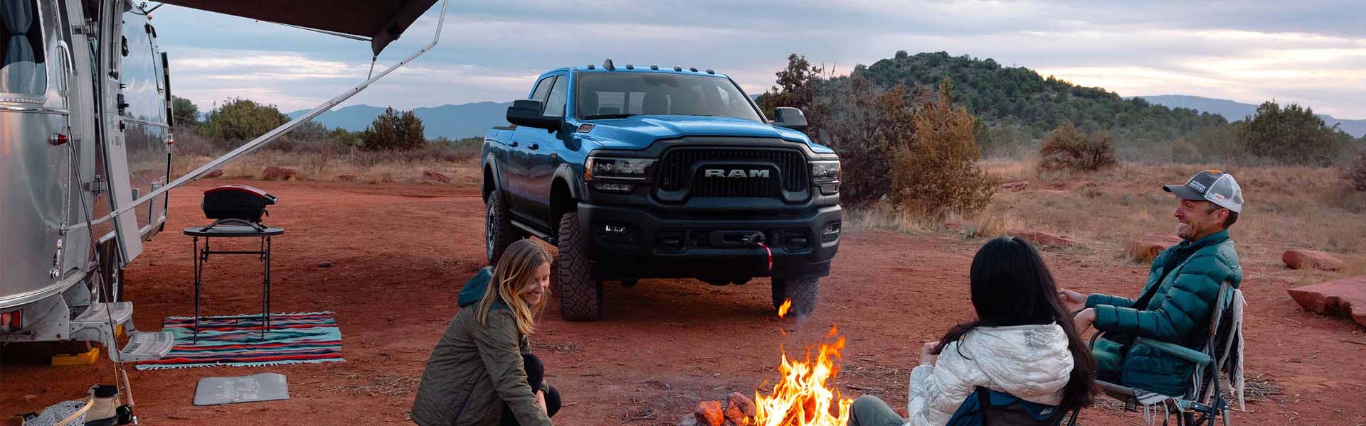 People around a campfire with a blue Ram truck next to an Airstream trailer in a desert landscape.