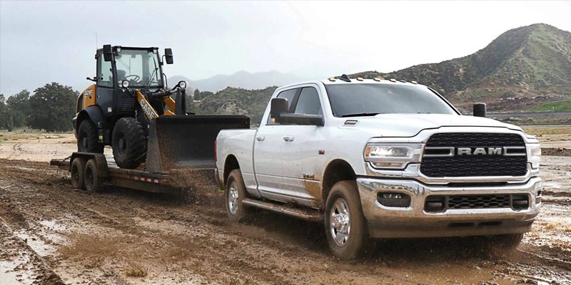A white Ram pickup truck, splattered with mud, tows a trailer with a yellow loader through a muddy field.