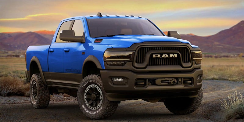 A blue Ram pickup truck with black trim, off-road tires, and a visible winch, parked on a dirt path with mountains in the background.