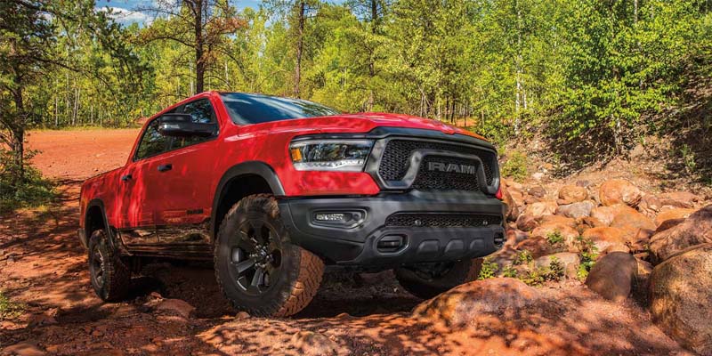 Red Ram pickup truck with black trim and off-road tires on a dirt trail with trees and rocks.