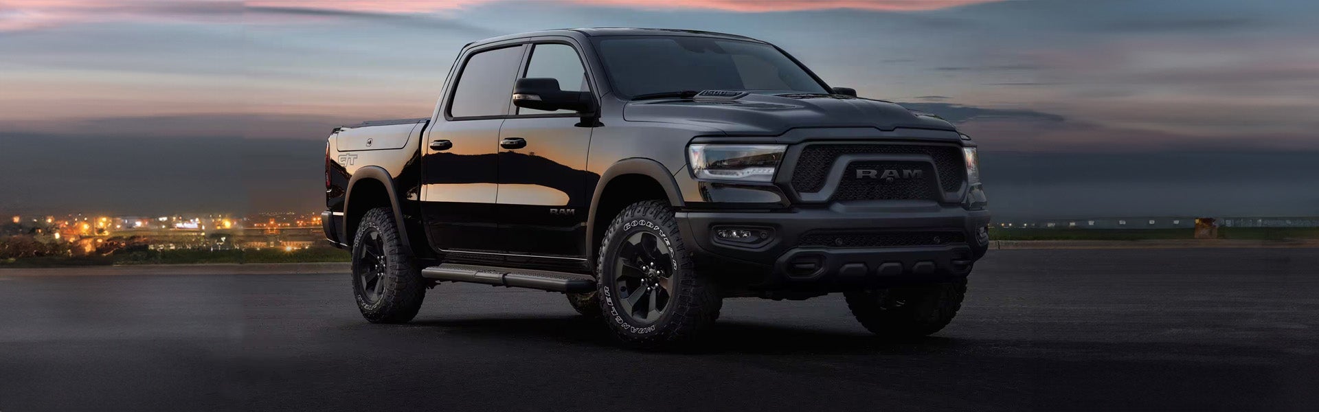 A black Ram pickup truck parked outdoors at dusk with city lights visible in the distant background.