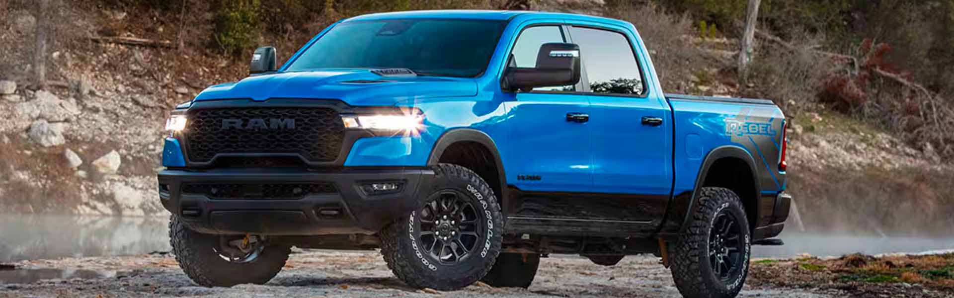 A bright blue Ram Rebel truck with shiny headlights sits on a dirt road next to water reflecting rocks and trees.