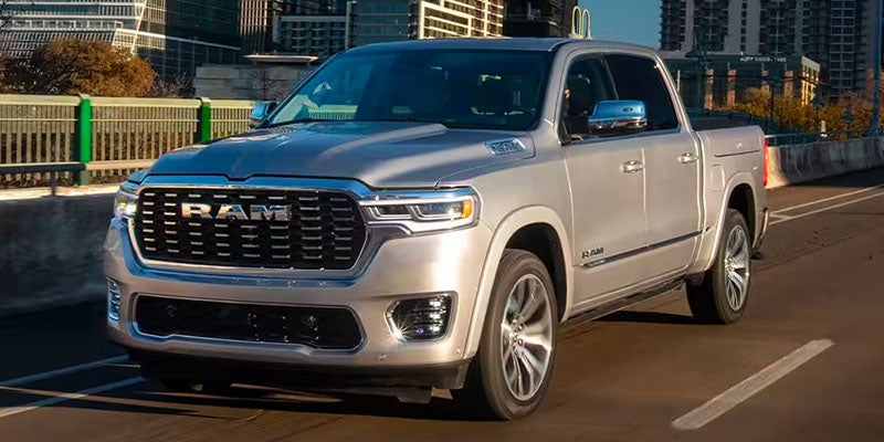 Silver 2025 Ram 1500 pickup truck driving on a multilane road with city buildings in the background.
