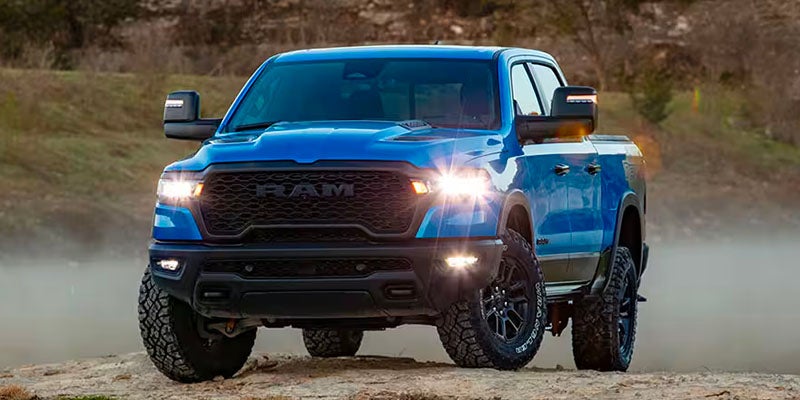 A bright blue Ram TRX pickup truck parked on dry, dusty terrain with its headlights illuminated.