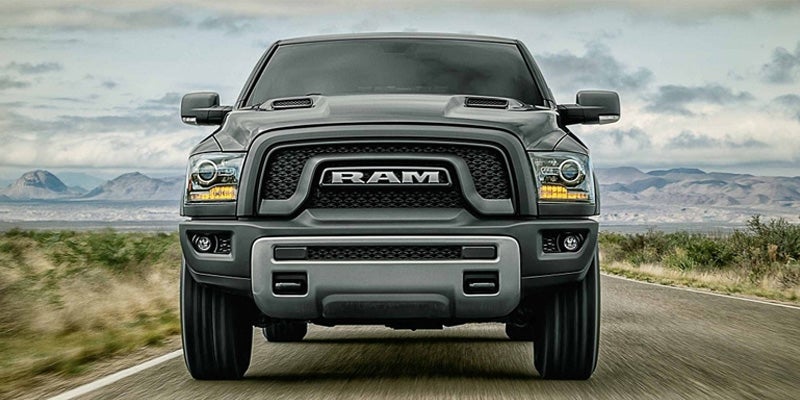 The front of a dark gray Ram pickup truck driving on a road, with mountains in the background.