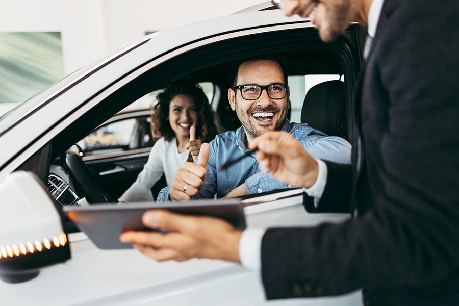 Happy couple in a new car giving thumbs up to a salesperson holding a tablet.